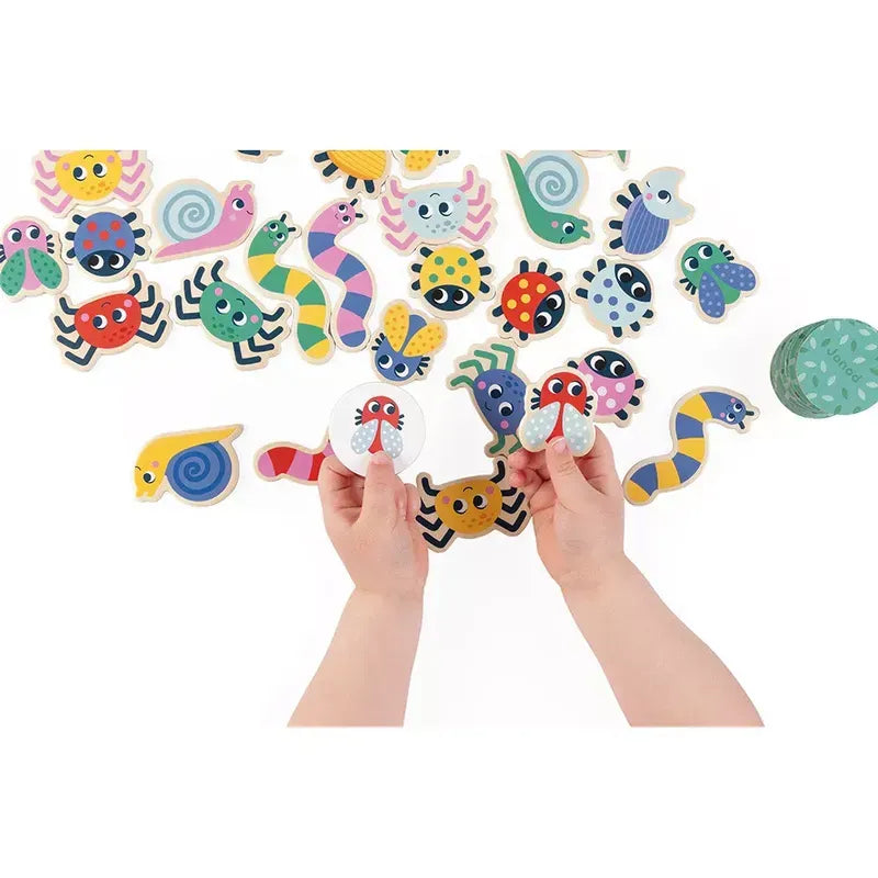 A pair of small hands holds two colorful wooden toys resembling bugs among an assortment of other brightly painted wooden insect toys, all part of the delightful Janod Seek & Find - The Garden game. The toys are spread out on a white surface, promoting observation through play.