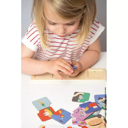 A young girl with blonde hair, wearing a white and red striped shirt, is deeply engrossed in the **Janod Sequences Cards - The Garden** educational toy at the table. She holds a puzzle piece as she works to enhance her logic and attention skills. Colorful pieces featuring various images of animals and objects are spread out before her.