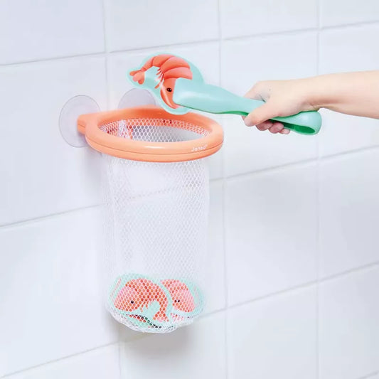 A child's hand placing a Janod Shrimp Catcher Bath Toy into a hanging mesh storage basket in a white tiled bathroom.