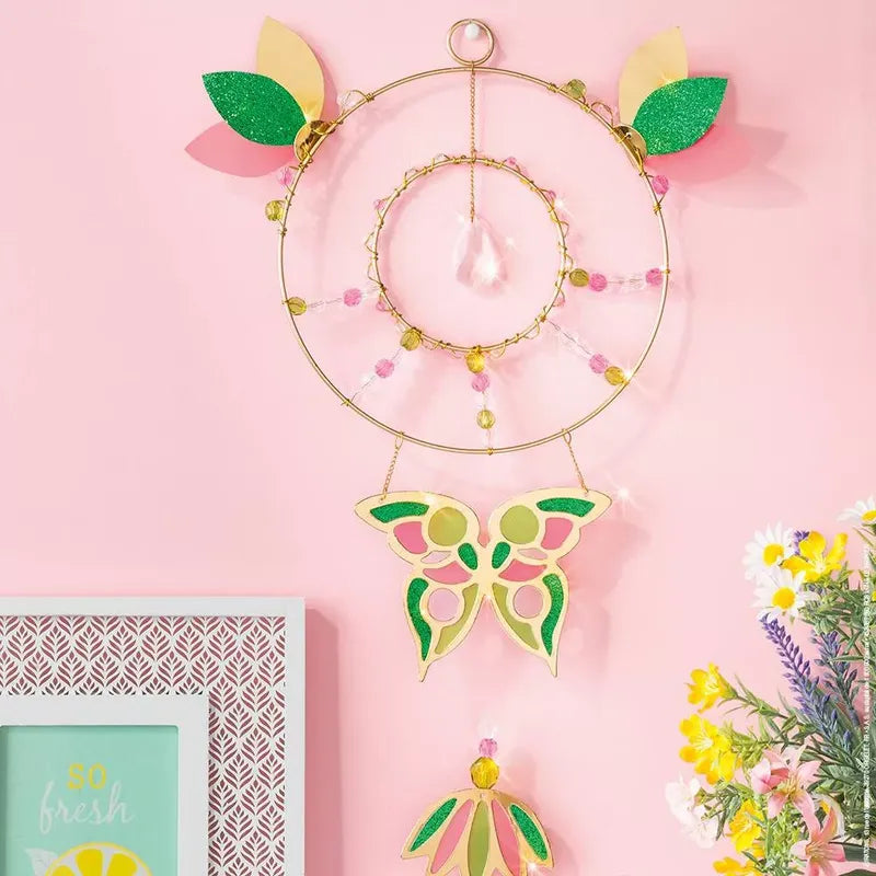 A Janod Suncatcher To Make, reminiscent of a children's craft kit, with a pink and yellow butterfly and green leaves hangs on a pink wall. Below it is a framed picture and a bouquet of colorful flowers. The suncatcher features beads, a crystal drop, and a smaller ring inside the larger one.