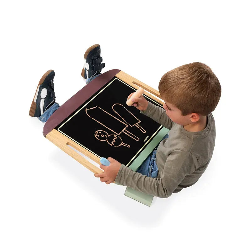 A child sits with the Janod White And Blackboard Table on their lap, using chalk to draw a popsicle and a small figure. The child is wearing a khaki shirt and blue jeans, focusing intently on their artwork. The table has handles on the sides for easy portability.