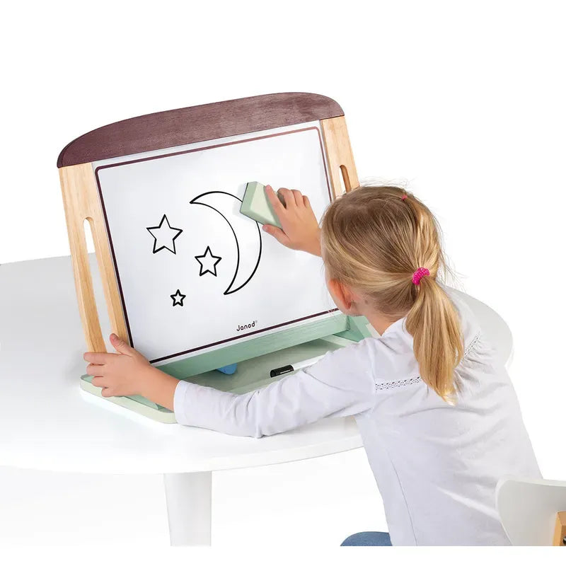 A young child with blonde hair tied in a ponytail is sitting at a table, erasing a drawing of a crescent moon and stars from the Janod White And Blackboard Table. The easel has a wooden frame and is placed on the white table. The child is wearing a white long-sleeved shirt.