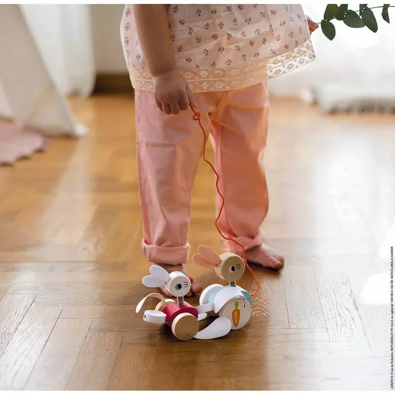 A little girl standing on a wooden floor, grasping the Janod Pull-along Rabbits and developing her motor skills.