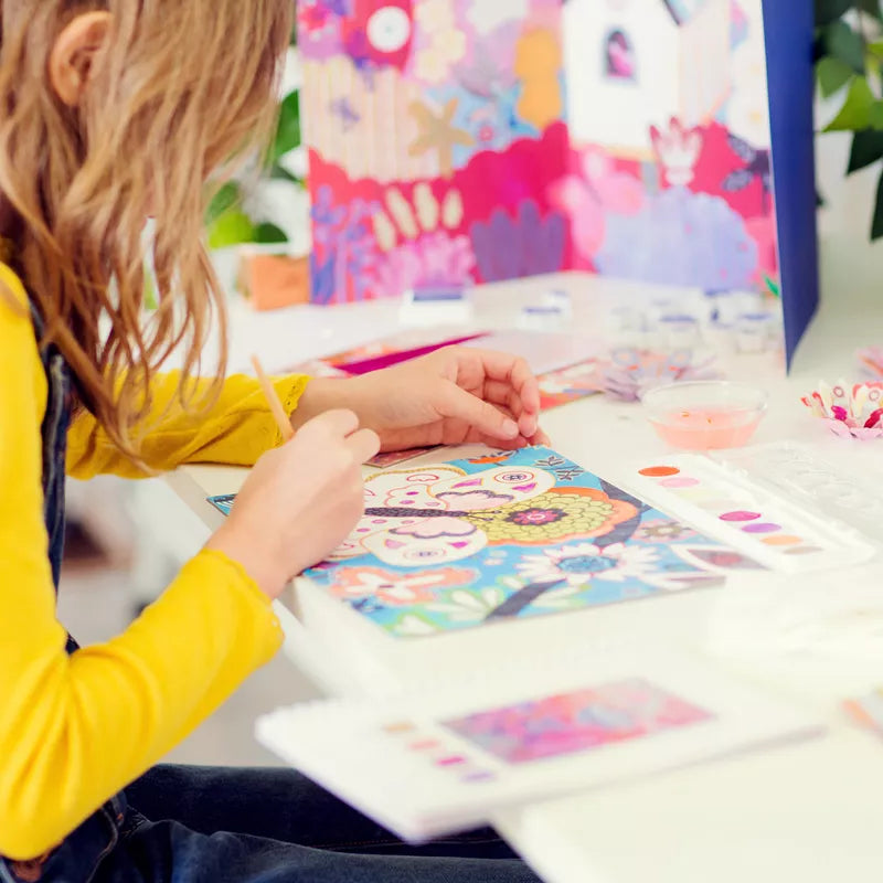 A little girl sitting at a table working on a painting with the Djeco Multi Activity Flower Box from Djeco.